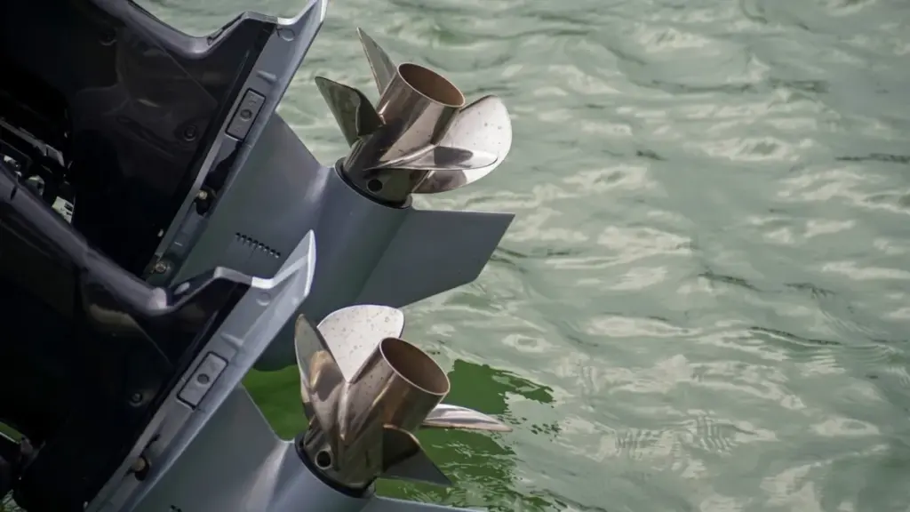 A close-up shot of twin stainless steel propellers on grey outboard boat motors positioned above slightly rippled, light green water