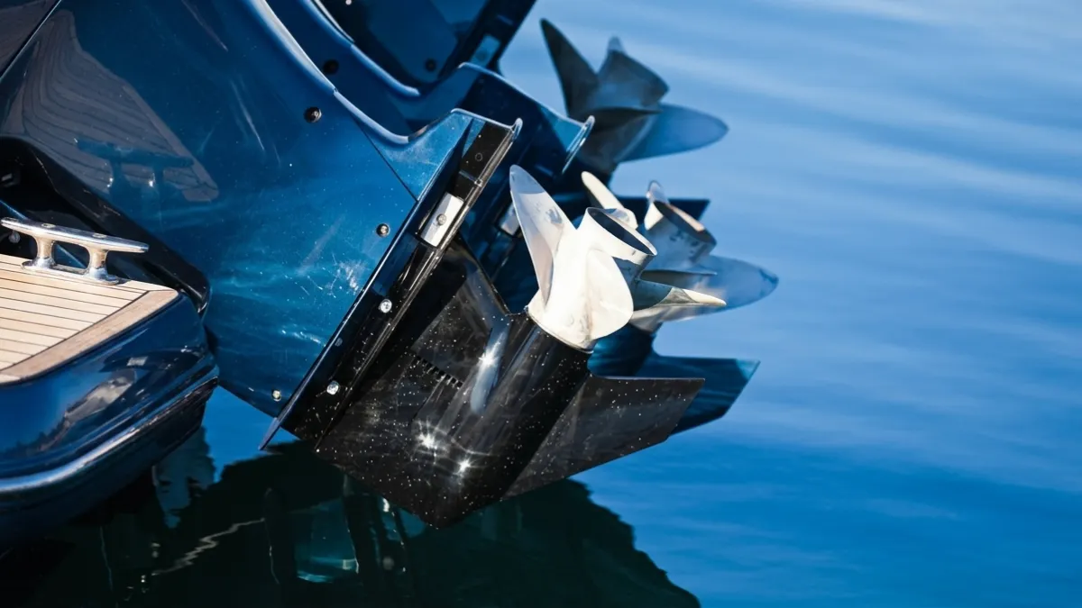 Uno scatto dettagliato delle eliche gemelle in acciaio inossidabile e delle unità inferiori di due motori fuoribordo blu, montato sullo specchio di poppa con ponte in teak, con acqua blu calma sullo sfondo