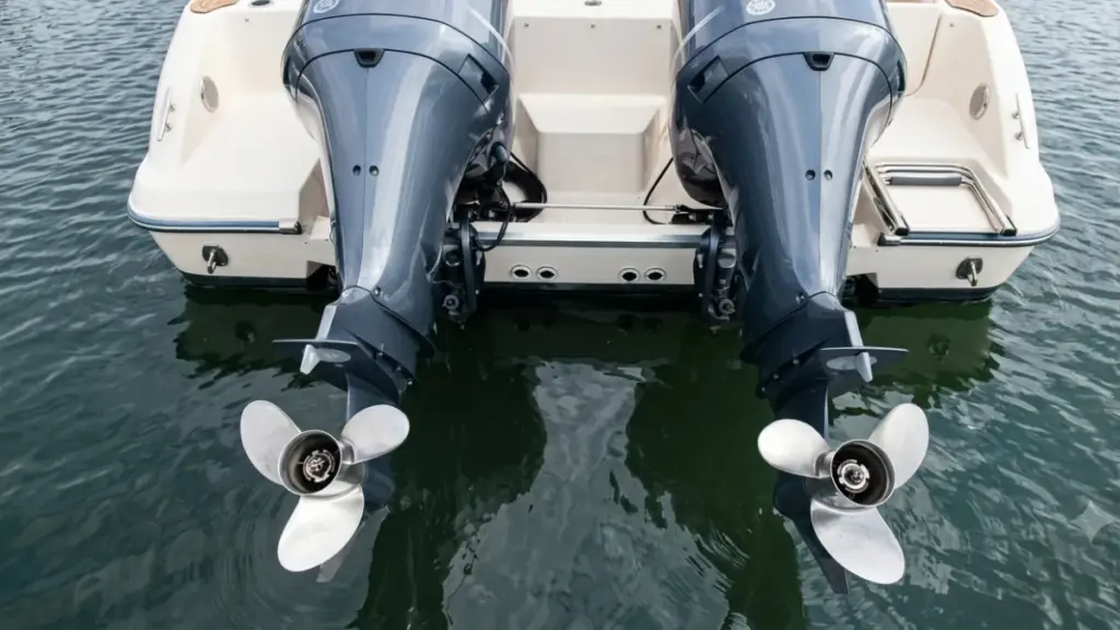 A high-angle, detailed shot of twin grey outboard boat motors with stainless steel propellers, mounted on the transom of a white boat over calm water.